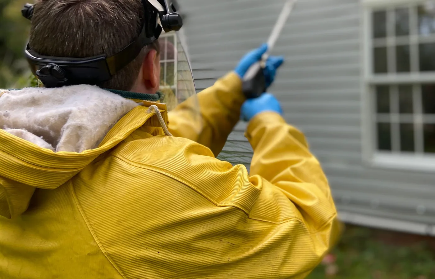 Technician in a yellow rain suit and face shield spraying the lap siding of a house