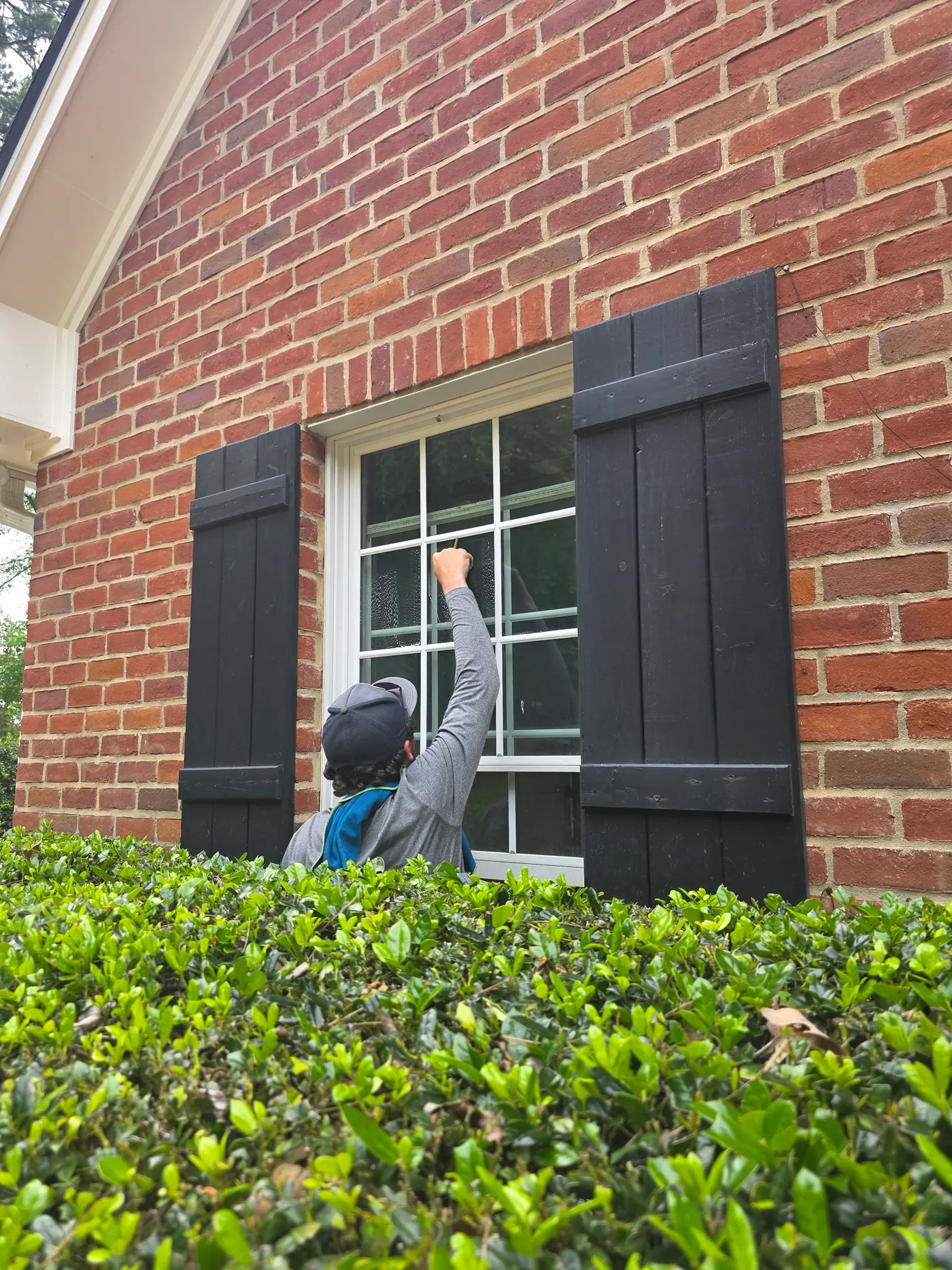 Technician cleaning a window on a brick home beside dark shutters
