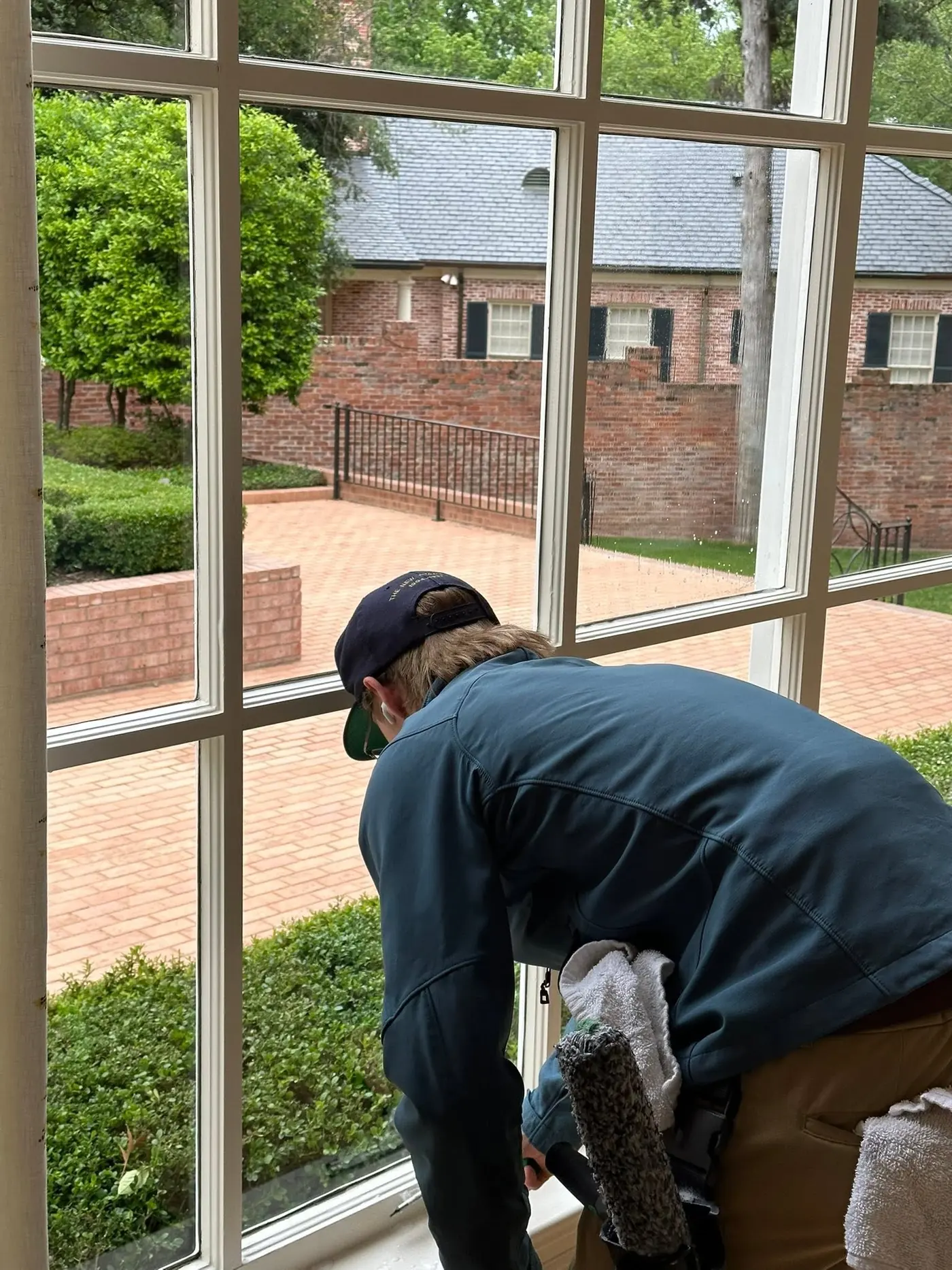 Technician wiping down a tall multi-pane window wall from indoors, a brick courtyard beyond the glass