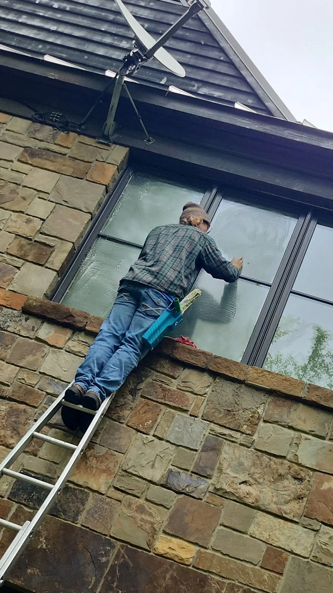 Technician on a ladder soaping a second-storey window on a stone wall, seen from the ground